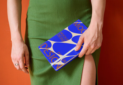 A notebook cover mockup is held in one hand by woman in a green skirt against an orange background