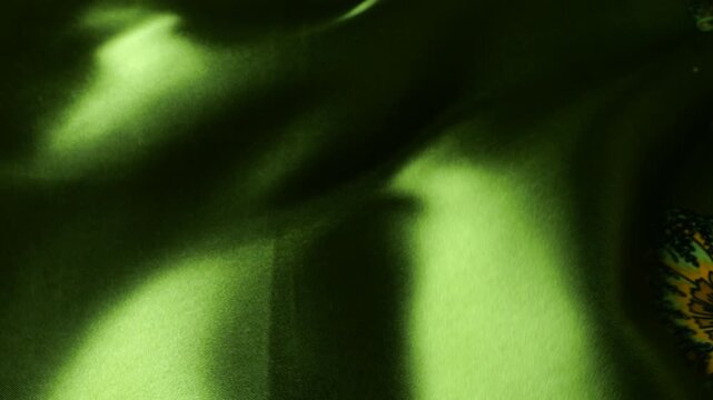 Close up of natural green silk fabric waving in the wind. Abstract background of moving soft waves on textile. Floral pattern with yellow and orange elements on dark green cloth.