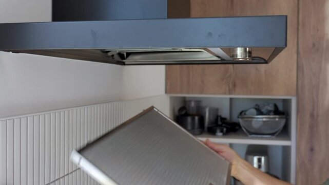 Hands removing metal grease filter from kitchen hood in home kitchen. Concept of appliance cleaning, maintenance and household care.
