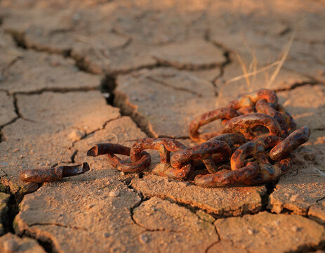 Rusty metal chain links lie on cracked, dry earth. The weathered surface and corroded metal represent drought and hardship. This scene evokes themes of perseverance and enduring strength.