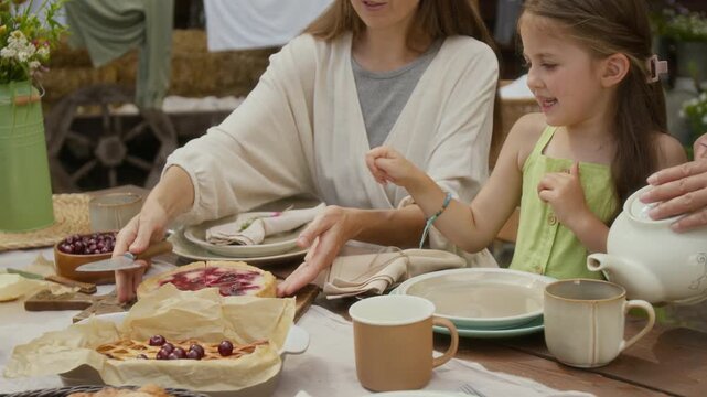 Medium shot of excited young 6-year-old Caucasian girl waiting for slice of homemade organic berry cheesecake while mother is cutting pie and father pouring tea, during family feast at rural farm