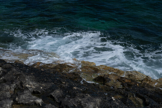 Waves crashing over rugged limestone rocks on the Maltese coastline, Mediterranean Sea surf creating white foam patterns against dark coastal stone. Natural seascape background with copy space.