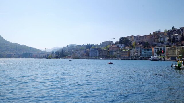 Wide panning shot of Lugano cityscape at lakefront with mountains and blue water, Switzerland