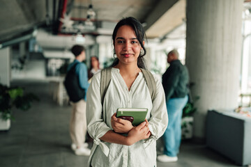 Naklejka na ściany i meble Young indian woman studying, holding books, wearing backpack, standing in a university hallway, looking at camera, smiling