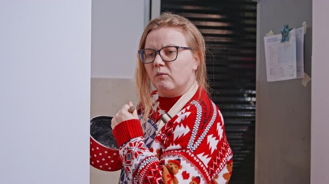A grown woman in a red sweater with a flour stained face peers out from around the corner holding a red saucepan with a whisk in the kitchen, looking away in surprise.