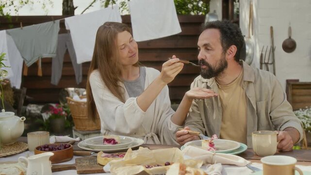 Medium shot of loving Caucasian rural wife feeding husband own homemade cheesecake, baked with new recipe, man enjoying and nodding with approval, while having lunch in yard in summer