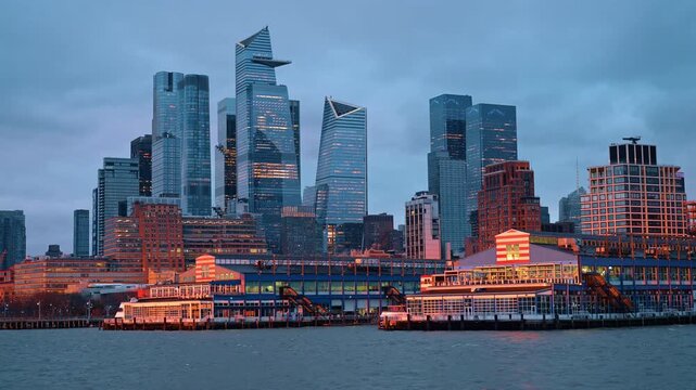 Hudson Yards skyline with Edge observation deck at golden hour. Waterfront view of Manhattan skyscrapers with warm sunset light hitting the facades and the Hudson River in foreground.