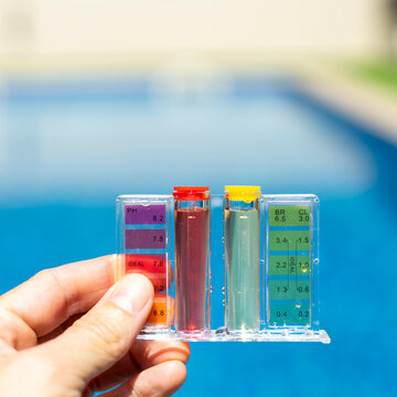 Close-up of a hand holding a pool water testing kit with color scale, measuring pH and chlorine levels in a clear blue swimming pool outdoors - pool maintenance concept