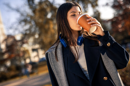 Young woman in autumn park holding takeaway coffee with headphones around neck