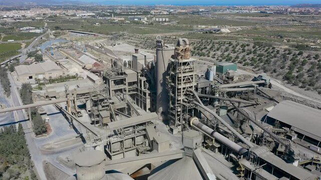 Aerial view of cement manufacturing plant in Alicante, Spain. Industrial complex with silos, conveyor systems and production facilities surrounded by agricultural landscape near Mediterranean coast. 