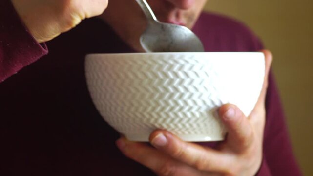 Close view of man eating ramen while holding bowl in one hand and spoon in another in natural home setting. Conveys authentic daily routine, solo eating moment, and unstyled lifestyle behavior.