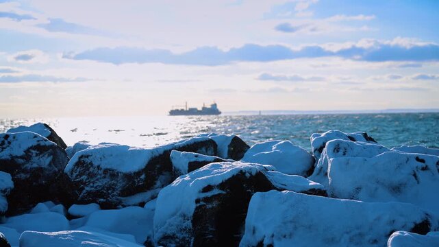 Snow covered rocks on breakwater with cargo ship in distance. Close view of icy boulders on a beach jetty with a freighter vessel on the sea horizon.