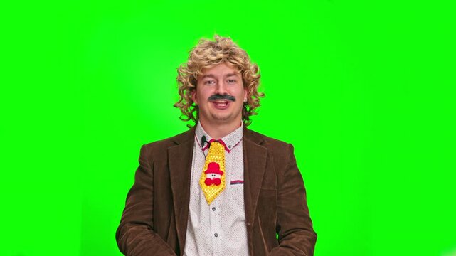 Mature man with curly blond wig and moustache wearing brown jacket and funny Christmas tie pointing at camera against green background, comic presenter concept, entertaining scene from studio life.