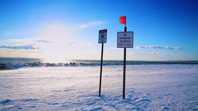 Warning signs and red flag on snow covered winter beach. No swimming and danger signs standing on a snowy beach under bright sunlight in New York City.