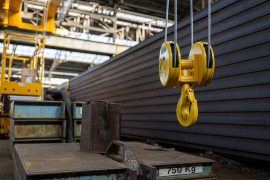 Industrial crane hook and pulley system hanging near stacked metal beams and counterweights inside large factory space showing heavy lifting and material handling environment
