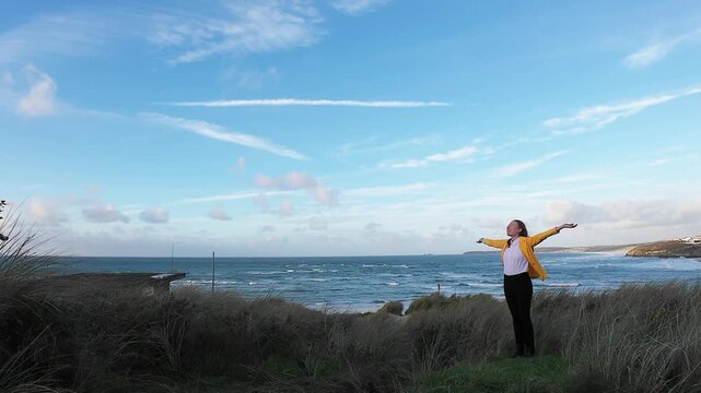 Relaxed Lady Stands On Rocky Coast. Outdoor Enthusiast Smiles Amidst Seaside Landscape And Sky