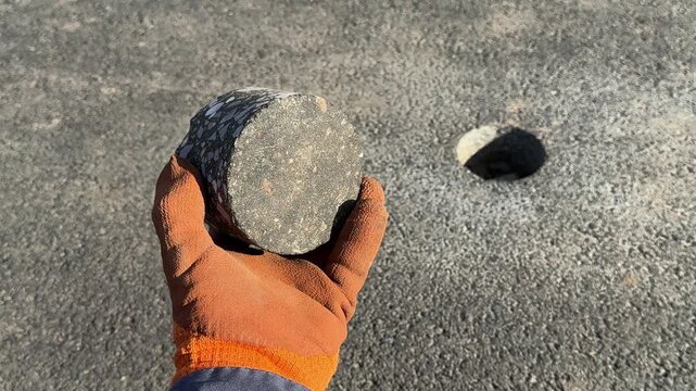 Asphalt core sample in gloved hand with drilled hole in road surface