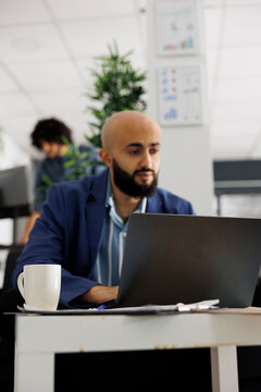 Arab project manager chating with coworker online on laptop while working in business office. Young financial analyst analyzing statistics report in corporate coworking space