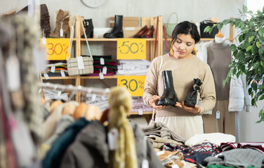 Fototapeta premium Latina woman in a warm sweater stands near the counter in a clothing store and chooses shoes with flat soles and heeled ankle boots. Woman buys ankle boots for autumn and winter
