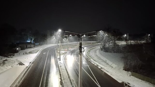 Snow drifts softly through the air as a car creeps along an icy road, its path illuminated by the glow of street lamps on a serene winter evening