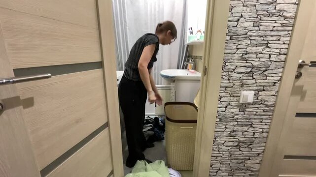Indoor scene of woman tidying garments. Domestic woman arranging clothing near wicker container inside hallway. Woman is diligently folding and smoothing green blanket inside narrow corridor