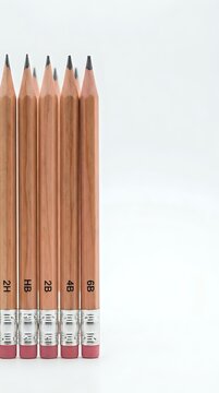 Five wooden pencils with pink erasers arranged in row showing different hardness grades from 2H to 6B on white background for office and school supplies.