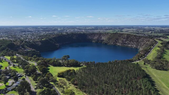 Blue Lake, Mount Gambier, South Australia in autumn: 4K aerial drone footage of volcanic crater lake, deep blue water, surrounding forest, cliffs, trails, seasonal foliage, farmlands, and houses