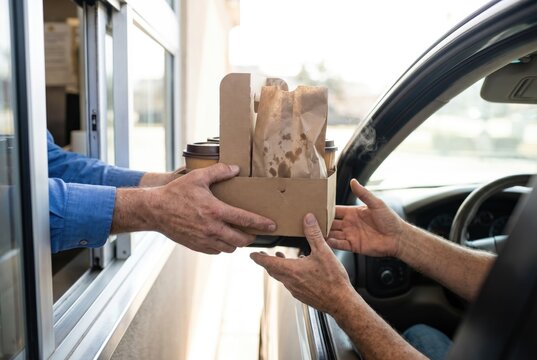 Worker handing paper tray with coffee cups and food bag to customer through drive thru window