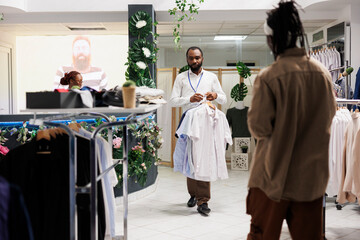 Fototapeta premium Clothes retail store african american man worker carrying shirts for customer to try. Buyer standing near garment rack and waiting for shop assistant to bring apparel different sizes