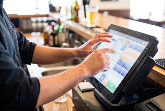 Bartender hands tapping drink order into wet electronic touchscreen POS system at bar counter