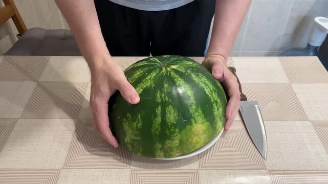 tabletop halved watermelon being sliced by white adult, hands guiding chef knife over striped rind, seeds visible, juicy red flesh, patterned tablecloth, plate and knife beside, cook demonstrating