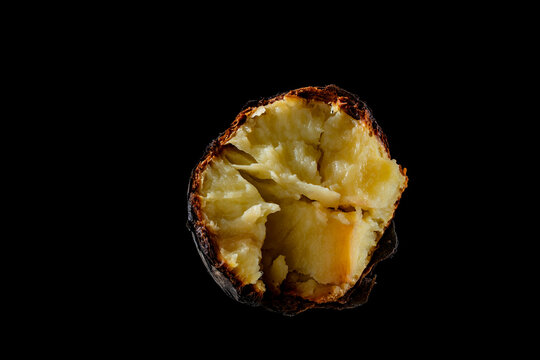Single baked potato in skin isolated on black background. Macro view of a whole potato baked in coals split open to show fluffy yellow texture. Rustic organic food concept isolated on black