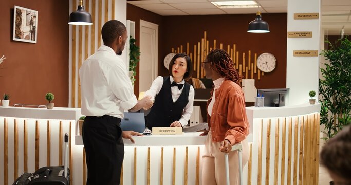 Friendly asian receptionist offering room key to happy guests after check in process is finalized. Busy exhausted hotel manager ensuring booking paperwork details are correct