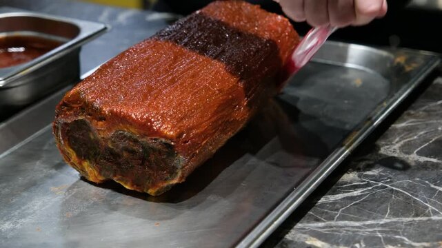 Action shot of a chef applying sauce to a large meat roll focusing on the texture of the marinade and the industrial feel of a professional restaurant kitchen during food prep.