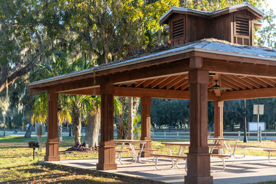 A large four-sided wooden gazebo overlooking a park with tall trees. The metal roof has a cedar wood cupola on top with louvred box vents. The structure has open sides and a concrete base floor. 