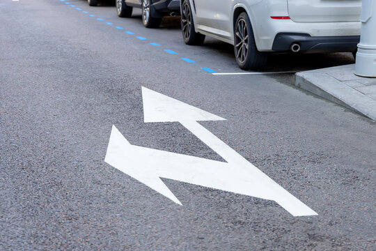 White road markings with two arrows on asphalt street near parked cars. Traffic direction signs and blue parking lines on urban road for city transport navigation.