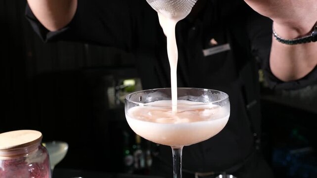 Expert bartender pouring a pink frothy cocktail into a coupe glass focusing on the smooth liquid texture and professional pouring technique for a luxury hospitality marketing campaign.