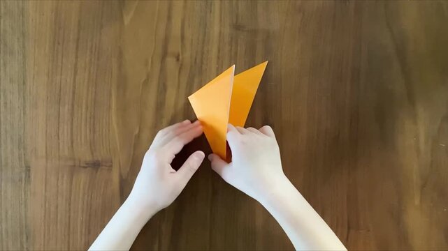 child hands folding orange origami paper on wooden table slow motion
