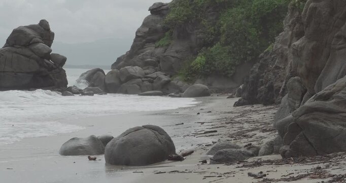 A rocky beach scene features large boulders along the shore with ocean waves crashing. Lush greenery is present on the cliffs in the background
