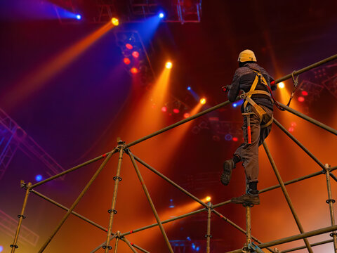 Stage construction for concerts, event production. Technician in safety harness climbs metal scaffolding installing lighting truss structures under colorful spotlights inside performance venue.