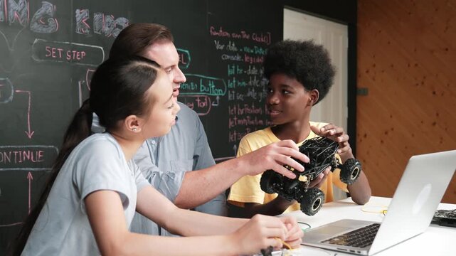 Smart teacher pick car model and give to happy student inspect robotic construction while sitting at table with electronic tool and laptop with display programming and coding programming. Edification
