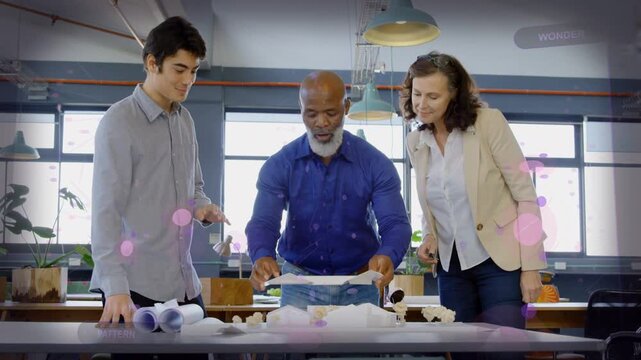 Design team leaning over plans, man in blue unfolding prototype for review, pink circles overlaying