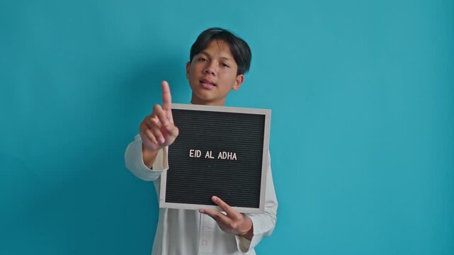 Muslim teenager holds "EID AL ADHA" letter board and wagging index finger in "no" or "don't" gesture on blue background