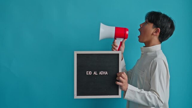 Side view Enthusiastic Muslim teenager announcing Islamic holiday through loudspeaker while holding "EID AL ADHA" sign isolated on blue background