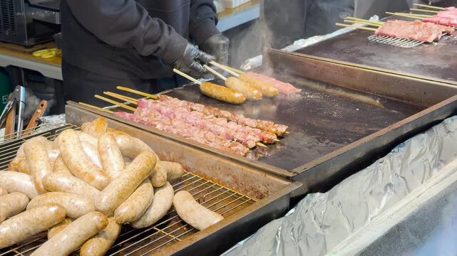 Grilling assorted meat skewers and sausages on flat top griddle at busy night market food stall.