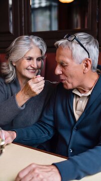 Playful senior wife teasingly persuading her focused husband to taste a new dish during an intimate restaurant date.