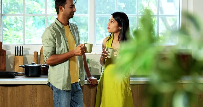 Indian asian young husband and wife having coffee at kitchen platform and surfing smartphone in joyful atmosphere in modern Indian home while sharing digital content and laughing during weekend