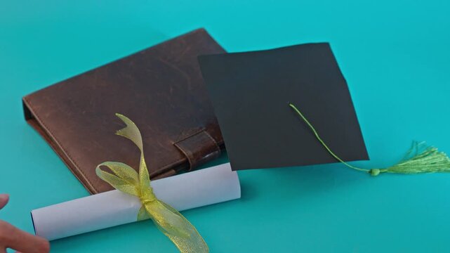 Hand placing diploma scroll with ribbon next to leather notebook and graduation cap on turquoise background