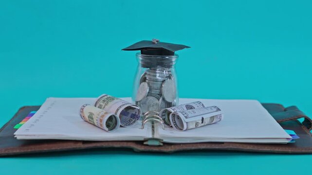 Glass jar filled with coins and topped with graduation cap on open notebook with rolled dollar bills