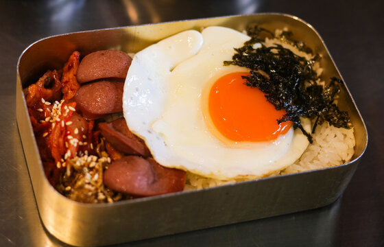 A traditional Korean dosirak served in a classic rectangular metal tin. The lunch box is packed with rice and topped with sunny-side-up egg, seaweed, sausages, kimchi and anchovies.
